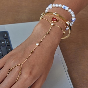 May include: A gold-toned hand chain featuring small heart-shaped charms and a central red heart accent, worn with a gold-toned bangle and a white beaded bracelet with blue and white patterned cylindrical beads on a wrist.