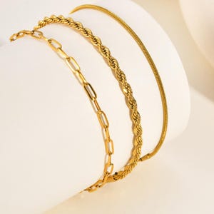 May include: A set of three gold-toned bracelets featuring distinct chain styles. The collection includes a delicate paperclip chain, a textured twisted rope chain, and a smooth, thin snake chain, all crafted with a polished metallic finish.