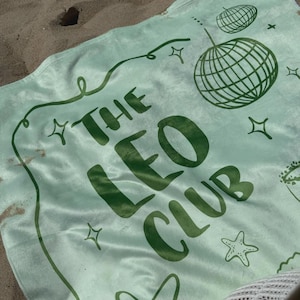 Pode incluir: Uma toalha de praia verde claro com o texto "THE LEO CLUB" em verde escuro. A toalha apresenta um design de bola de discoteca, estrelas e uma estrela do mar. A toalha est&aacute; na areia.