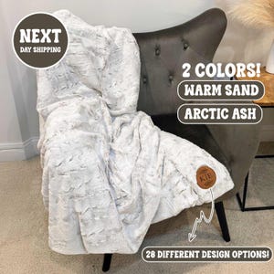 May include: A plush, textured blanket in a neutral color draped over a gray armchair. The blanket has a soft, fur-like appearance. The image includes text that reads "2 Colors! Warm Sand, Arctic Ash" and "28 Different Design Options!"