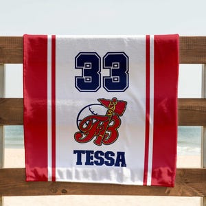 May include: A white and red baseball towel with the number 33 in navy blue. The towel features a baseball and axe logo with the name "Tessa" in navy blue. The towel is hanging on a wooden fence with a beach and ocean in the background.