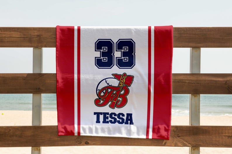 May include: A white and red baseball towel with the number 33 in navy blue. The towel features a baseball and axe logo with the name "Tessa" in navy blue. The towel is hanging on a wooden fence with a beach and ocean in the background.