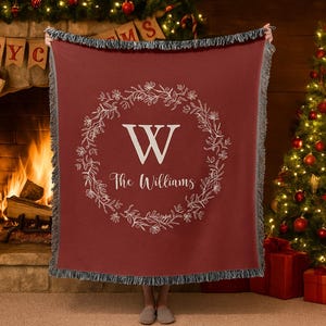 May include: A burgundy throw blanket with a white floral wreath design and the letter "W" in the center. The words "The Williams" are written below the letter. The blanket has a fringed edge and is displayed in front of a fireplace and Christmas tree.