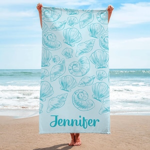 Puede incluir: Una toalla de playa azul claro con un estampado de conchas marinas turquesas. El nombre "Jennifer" est&aacute; impreso en una fuente cursiva en la parte inferior. La toalla se muestra sobre un fondo de playa y oc&eacute;ano, con el cielo visible.