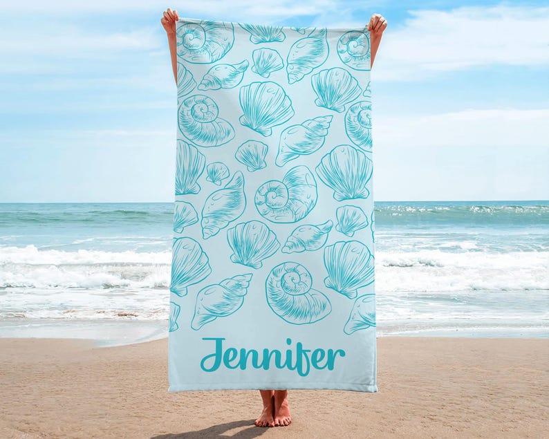 Puede incluir: Una toalla de playa azul claro con un estampado de conchas marinas turquesas. El nombre "Jennifer" est&aacute; impreso en una fuente cursiva en la parte inferior. La toalla se muestra sobre un fondo de playa y oc&eacute;ano, con el cielo visible.