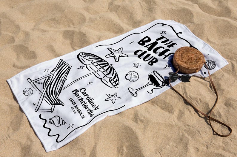 Pode incluir: Toalha de praia branca com ilustra&ccedil;&otilde;es pretas de uma cadeira de praia, guarda-sol, conchas e um copo de cocktail. A toalha diz "The Bach Club" e "Caroline's Bachelorette Santa Barbara, CA EST. 2015". Uma bolsa redonda de tecido e &oacute;culos de sol est&atilde;o na areia.