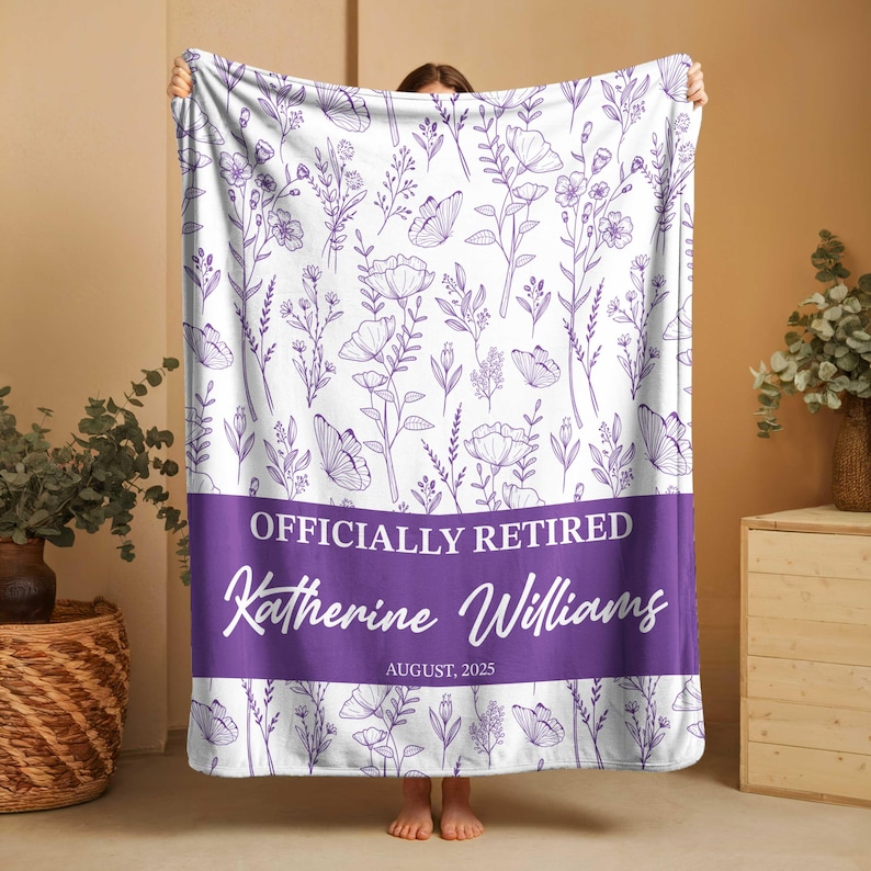 May include: A white blanket featuring a purple floral and butterfly design. The blanket has a purple band with the text "OFFICIALLY RETIRED" and the name "Katherine Williams" with the date "AUGUST, 2025".