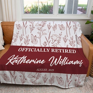 May include: A white and claret throw blanket featuring a floral and butterfly design. The blanket has a claret banner with the text "OFFICIALLY RETIRED" and the name "Katherine Williams" with the date "AUGUST, 2025".