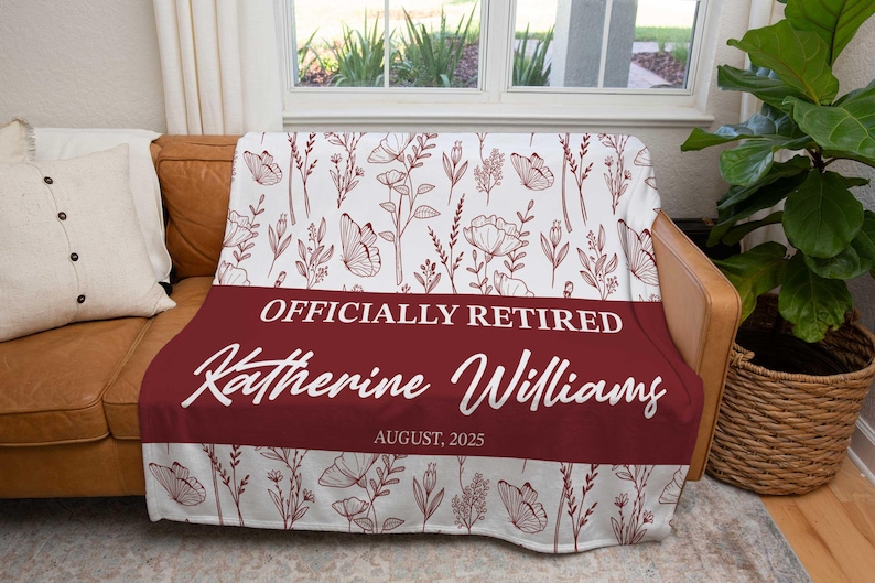 May include: A white and claret throw blanket featuring a floral and butterfly design. The blanket has a claret banner with the text "OFFICIALLY RETIRED" and the name "Katherine Williams" with the date "AUGUST, 2025".