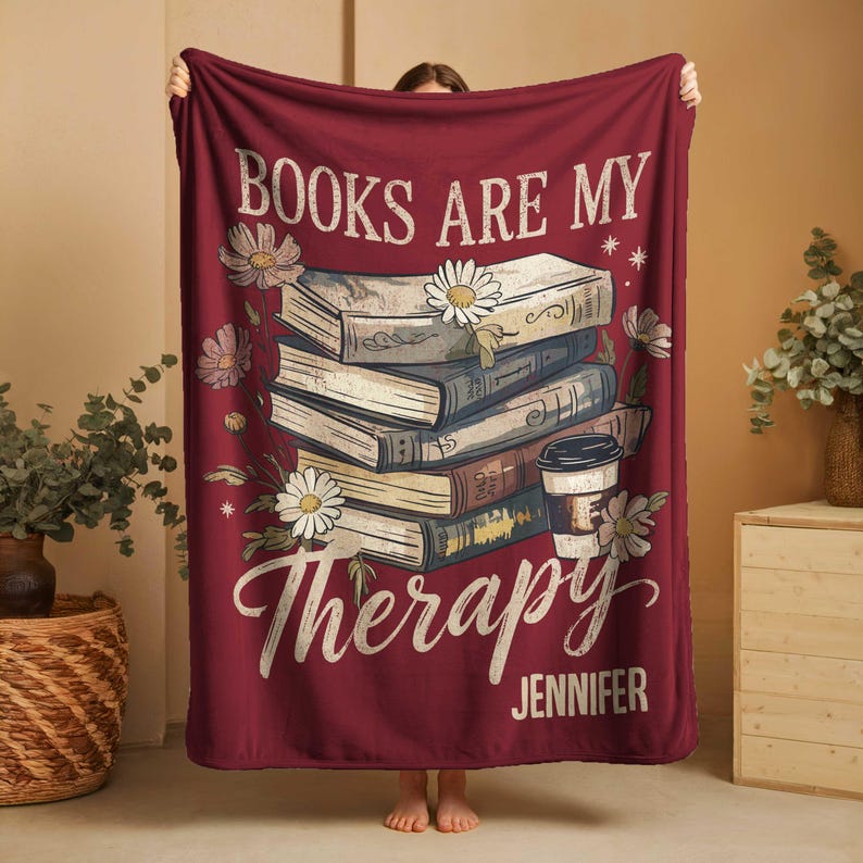 Peut inclure: Une couverture bordeaux avec le texte "BOOKS ARE MY Therapy", une pile de livres, des marguerites et une illustration de tasse &agrave; caf&eacute;. Le nom "Jennifer" est imprim&eacute; en bas. La couverture est tenue par une personne.