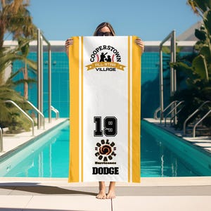 May include: A white and yellow beach towel featuring the text "COOPERSTOWN ALL STAR VILLAGE" and "19 GOLD COAST HURRICANES DODGE". The towel is held in front of a swimming pool.