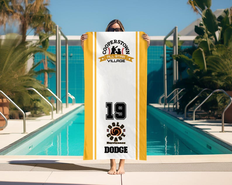 May include: A white and yellow beach towel featuring the text "COOPERSTOWN ALL STAR VILLAGE" and "19 GOLD COAST HURRICANES DODGE". The towel is held in front of a swimming pool.