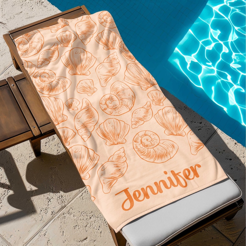 Puede incluir: Toalla de playa color melocot&oacute;n con un patr&oacute;n repetido de ilustraciones de conchas marinas naranjas. La toalla est&aacute; personalizada con el nombre "Jennifer" en escritura naranja. La toalla est&aacute; sobre una tumbona de madera junto a una piscina.