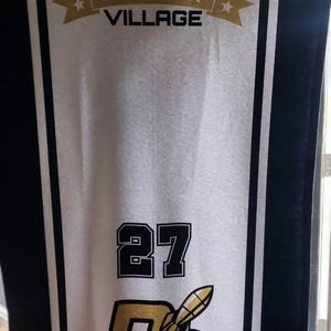 May include: A white banner with black and gold detailing, displaying the text "COOPERSTOWN ALL STAR VILLAGE." The banner also shows the number "27" and the letters "DC" with a baseball bat graphic, and the name "LENDORF."