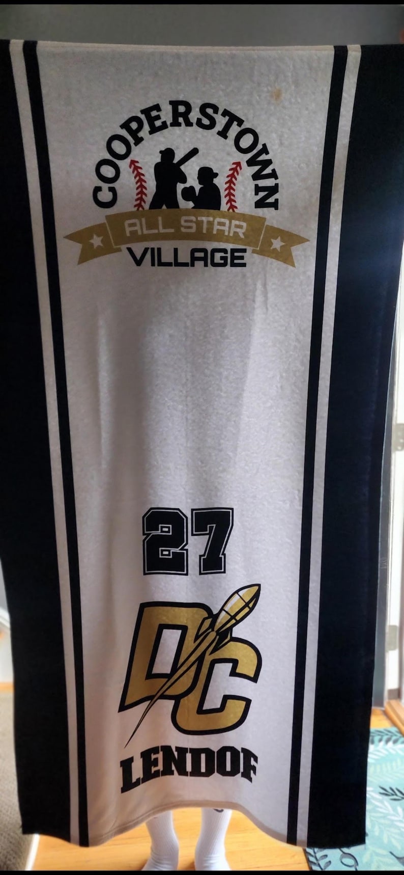 May include: A white banner with black and gold detailing, displaying the text "COOPERSTOWN ALL STAR VILLAGE." The banner also shows the number "27" and the letters "DC" with a baseball bat graphic, and the name "LENDORF."