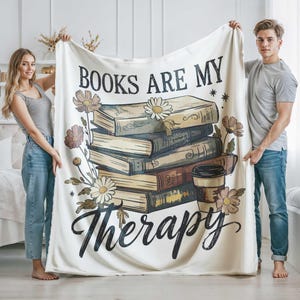 Peut inclure: Plaid de couleur cr&egrave;me avec l'inscription "BOOKS ARE MY Therapy" en &eacute;criture noire. La couverture pr&eacute;sente une pile de livres, des fleurs et un dessin de tasse &agrave; caf&eacute;. La couverture est tenue par deux personnes.