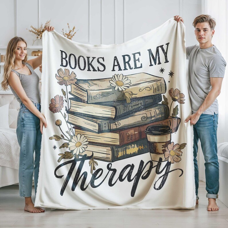 Peut inclure: Plaid de couleur cr&egrave;me avec l'inscription "BOOKS ARE MY Therapy" en &eacute;criture noire. La couverture pr&eacute;sente une pile de livres, des fleurs et un dessin de tasse &agrave; caf&eacute;. La couverture est tenue par deux personnes.