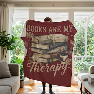 Peut inclure: Une couverture bordeaux avec l'inscription "BOOKS ARE MY Therapy" et un motif de livres, de marguerites et d'une tasse de caf&eacute;. La couverture est pr&eacute;sent&eacute;e, mettant en valeur sa texture douce et son design d&eacute;coratif.