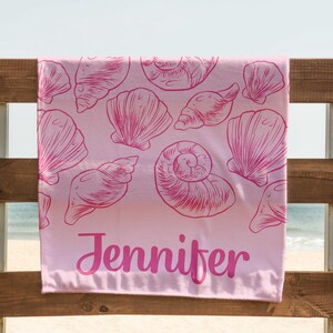 Puede incluir: Una toalla de playa rosa con un dise&ntilde;o de conchas marinas y el nombre "Jennifer" en cursiva. La toalla est&aacute; colgada de una valla de madera, con el mar visible al fondo.
