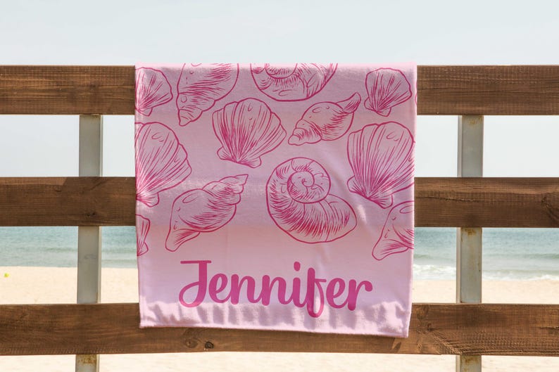 Puede incluir: Una toalla de playa rosa con un dise&ntilde;o de conchas marinas y el nombre "Jennifer" en cursiva. La toalla est&aacute; colgada de una valla de madera, con el mar visible al fondo.