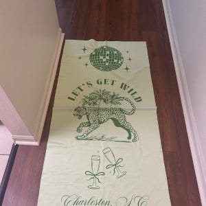 Pode incluir: Bandeira de tecido verde claro com uma bola de discoteca, estrelas e uma ilustra&ccedil;&atilde;o de leopardo. O texto "LET'S GET WILD" est&aacute; acima do leopardo. Duas ta&ccedil;as de champanhe est&atilde;o abaixo, com o texto "Charleston, SC" na parte inferior.