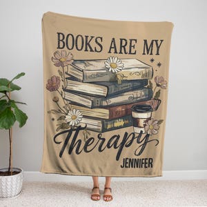 Peut inclure: Plaid beige avec l'inscription "BOOKS ARE MY Therapy" et le nom "Jennifer". Le plaid pr&eacute;sente une pile de livres, des fleurs et une illustration de tasse &agrave; caf&eacute;. Un &eacute;l&eacute;ment de d&eacute;coration int&eacute;rieure confortable.