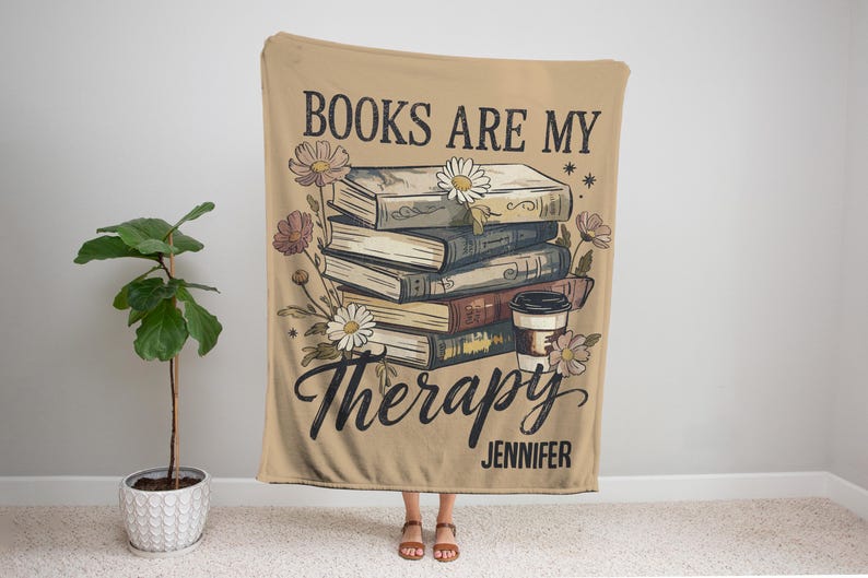 Peut inclure: Plaid beige avec l'inscription "BOOKS ARE MY Therapy" et le nom "Jennifer". Le plaid pr&eacute;sente une pile de livres, des fleurs et une illustration de tasse &agrave; caf&eacute;. Un &eacute;l&eacute;ment de d&eacute;coration int&eacute;rieure confortable.
