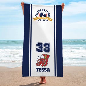 May include: A white beach towel with navy blue borders and stripes. The towel features the text "COOPERSTOWN ALL STAR VILLAGE" at the top, the number "33", and the name "TESSA" with a baseball graphic. The towel is held up on a beach.