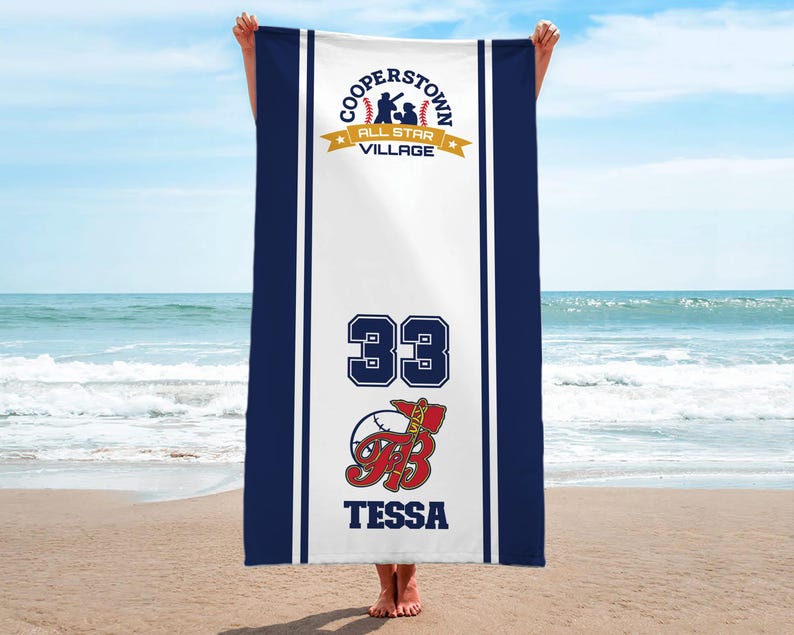 May include: A white beach towel with navy blue borders and stripes. The towel features the text "COOPERSTOWN ALL STAR VILLAGE" at the top, the number "33", and the name "TESSA" with a baseball graphic. The towel is held up on a beach.