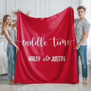 May include: A red blanket with the words "cuddle time" in white script. Below the text are the names "Hailey" and "Justin" with a heart symbol. The blanket is held up by a woman and a man.