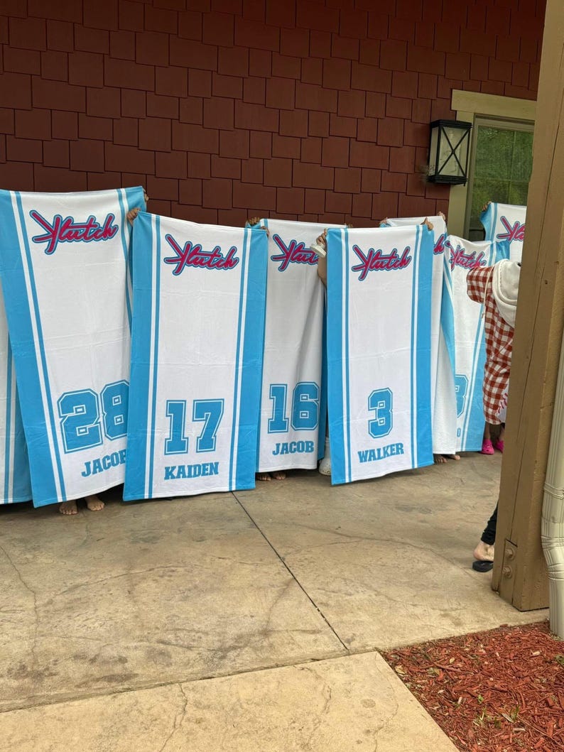 May include: A collection of white beach towels with blue borders, each bearing the word "Yatch" in a decorative script. The towels are personalized with numbers and names such as "28 JACOB", "17 KAIDEN", "16 JACOB", and "3 WALKER".