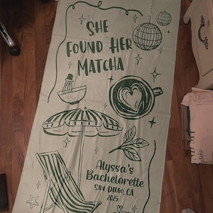 Pode incluir: Uma toalha de praia verde claro com tema matcha. A toalha apresenta o texto "SHE FOUND HER *MATCHA" e ilustra&ccedil;&otilde;es de um batedor de matcha, uma x&iacute;cara de caf&eacute; com um cora&ccedil;&atilde;o, um guarda-sol e uma cadeira de praia. O texto "Alyssa's Bachelorette SAN DIEGO, CA 2025" tamb&eacute;m est&aacute; impresso.
