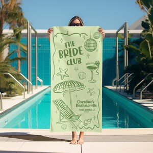 Pode incluir: Uma toalha de praia verde claro com o texto "The Bride Club" e ilustra&ccedil;&otilde;es com tema de praia. A toalha apresenta uma bola de discoteca, um cocktail, uma estrela do mar, uma concha e um guarda-sol. O texto "Caroline's Bachelorette" tamb&eacute;m &eacute; vis&iacute;vel.