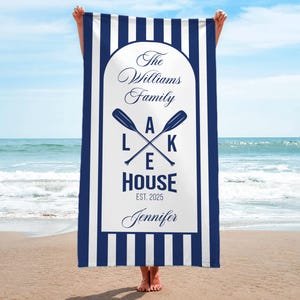 May include: A navy blue and white striped beach towel with a central white panel. The panel features the text "The Williams Family Lake House Est. 2025 Jennifer" with crossed oars. The towel is held up on a beach.