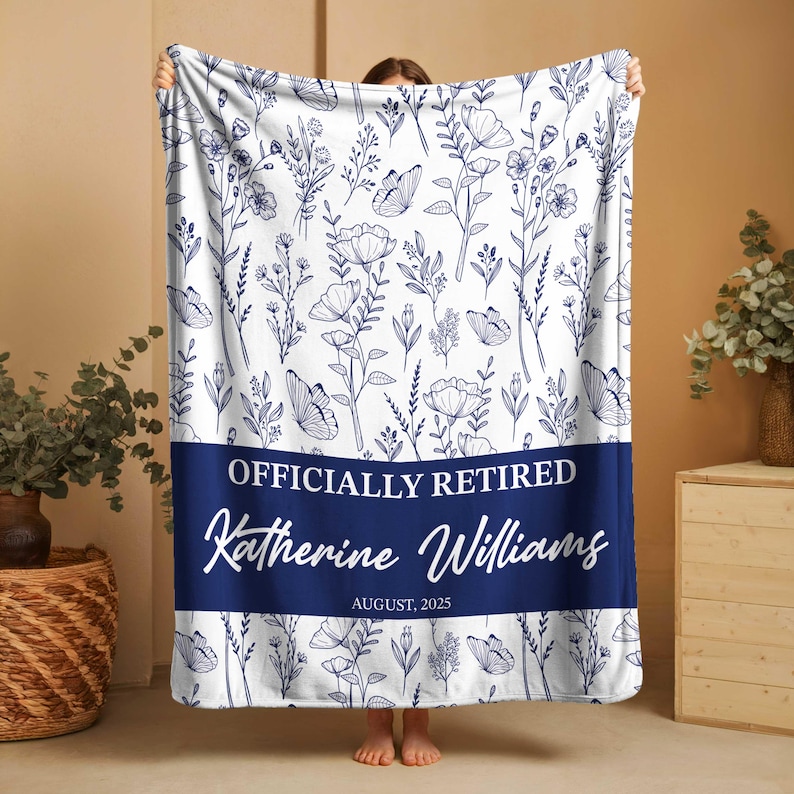 May include: A white blanket featuring a navy blue floral design and butterflies. The blanket has a navy blue band with the text "OFFICIALLY RETIRED", the name "Katherine Williams" and the date "AUGUST, 2025".
