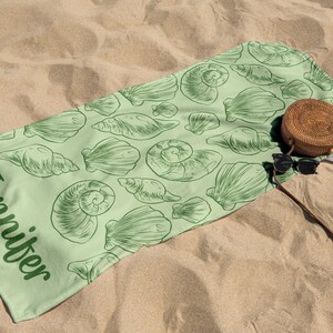 Puede incluir: Una toalla de playa verde claro con un dise&ntilde;o de conchas marinas y el nombre "Jennifer" impreso en verde. Una bolsa redonda tejida y gafas de sol descansan sobre la toalla, sobre arena.