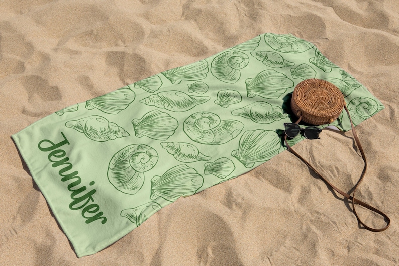 Puede incluir: Una toalla de playa verde claro con un dise&ntilde;o de conchas marinas y el nombre "Jennifer" impreso en verde. Una bolsa redonda tejida y gafas de sol descansan sobre la toalla, sobre arena.