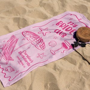 Pode incluir: Uma toalha de praia rosa com o texto "The Bride Club" e ilustra&ccedil;&otilde;es de uma cadeira de praia, um guarda-sol e conchas. Uma bolsa redonda de vime e &oacute;culos de sol repousam sobre a toalha, numa praia arenosa.