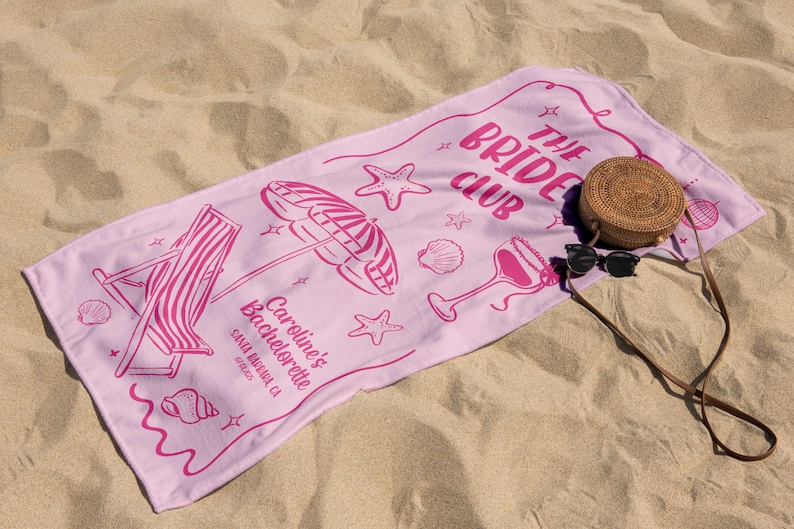 Pode incluir: Uma toalha de praia rosa com o texto "The Bride Club" e ilustra&ccedil;&otilde;es de uma cadeira de praia, um guarda-sol e conchas. Uma bolsa redonda de vime e &oacute;culos de sol repousam sobre a toalha, numa praia arenosa.