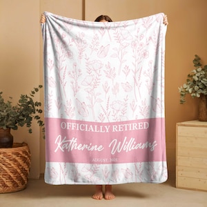 May include: A white and pink floral blanket featuring the text "OFFICIALLY RETIRED" and "Katherine Williams" in cursive. A pink band at the bottom displays the date "AUGUST, 2025". The blanket is held up by a person, showcasing its design.