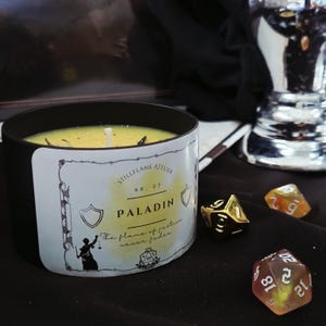 Op de afbeelding: Een zwarte kaars met een gele was binnenkant en een wit etiket met de tekst "PALADIN". De kaars staat naast drie dobbelstenen, één goudkleurig en twee doorschijnend met zichtbare cijfers. De tekst "The flame of justice never fades" is ook zichtbaar.