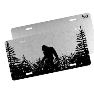 May include: A black and white metal license plate featuring a silhouette of Bigfoot walking through a forest. The words "Front" and "Back" are printed on the plate.