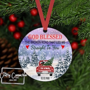 May include: A round Christmas ornament with a red ribbon. The ornament features a winter scene with a red truck carrying a Christmas tree. Text includes "GOD BLESSED", "Straight To You", names, and a date.