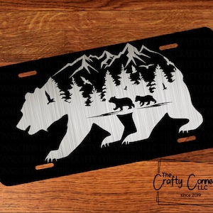 May include: A black metal license plate with a silver bear silhouette. The bear is walking through a mountain range with trees and two smaller bears behind it. The text "Crafty Connection LLC since 2019" is at the bottom of the plate.