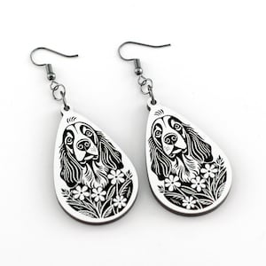 May include: A pair of white teardrop earrings featuring a black and white illustration of a dog's face with floral accents. The earrings have silver-colored hooks and are displayed against a white background.