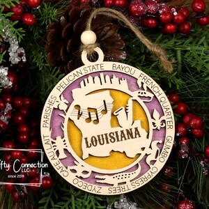 Puede incluir: Un adorno navide&ntilde;o de madera con forma de c&iacute;rculo con fondo morado y amarillo. El adorno presenta una silueta del estado de Luisiana con la palabra "Louisiana" escrita en su interior. El adorno est&aacute; rodeado de palabras que describen Luisiana, como "Bayou", "French Quarter", "Gumbo", "Cypress Trees", "Pelican State" y "Cafe au Lait".