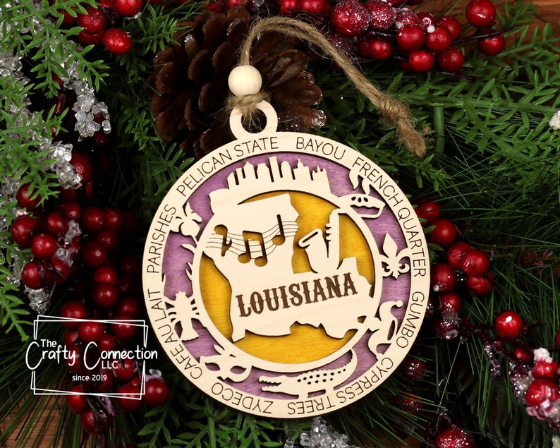 Puede incluir: Un adorno navide&ntilde;o de madera con forma de c&iacute;rculo con fondo morado y amarillo. El adorno presenta una silueta del estado de Luisiana con la palabra "Louisiana" escrita en su interior. El adorno est&aacute; rodeado de palabras que describen Luisiana, como "Bayou", "French Quarter", "Gumbo", "Cypress Trees", "Pelican State" y "Cafe au Lait".