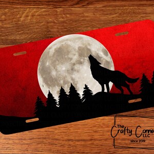 May include: License plate featuring a silhouette of a howling wolf against a red sky with a full moon. Black trees are in the foreground. The Crafty Connection LLC logo is in the bottom right corner.