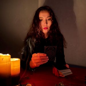 Pode incluir: Uma pessoa de cabelos escuros longos segura uma carta de tar&ocirc; retangular com um design geom&eacute;trico preto e dourado. Duas velas grossas acesas emitem fuma&ccedil;a, e um baralho de cartas repousa sobre uma superf&iacute;cie vermelha junto a ervas secas.