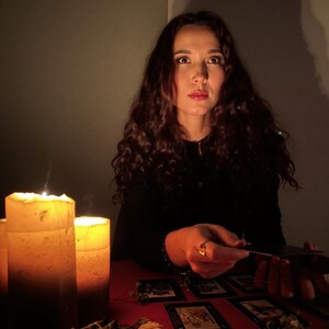 Puede incluir: Una mujer con largo cabello casta&ntilde;o rizado est&aacute; sentada en una mesa roja, iluminada por velas. Hay cartas del tarot dispuestas, y ella sostiene una. Un manojo de salvia y flores secas tambi&eacute;n est&aacute;n sobre la mesa, creando una atm&oacute;sfera m&iacute;stica.